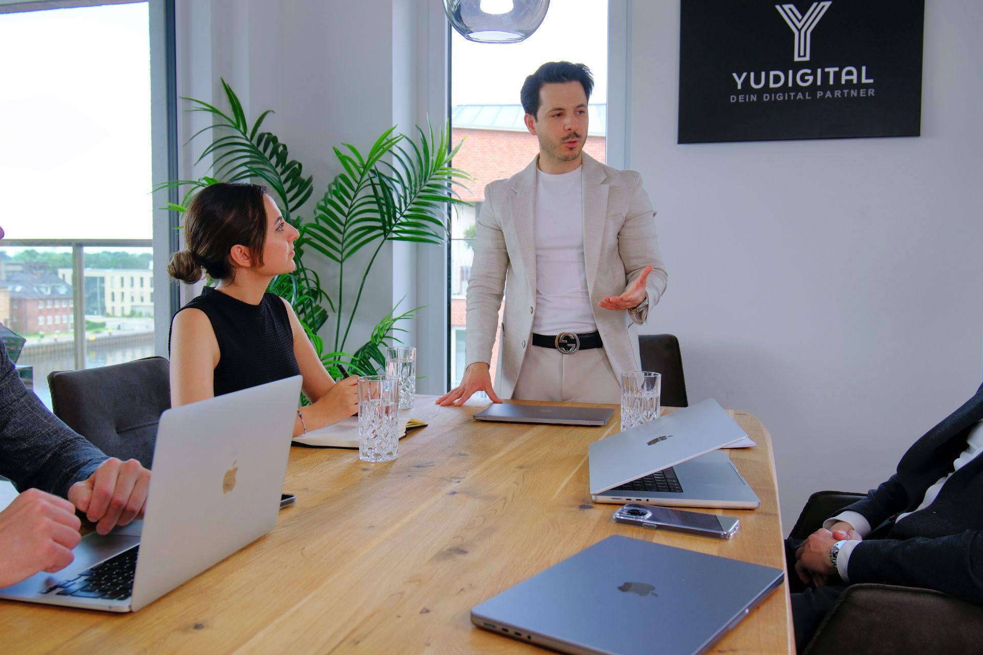 Business meeting with a man standing and talking to seated colleagues around a wooden table with laptops.